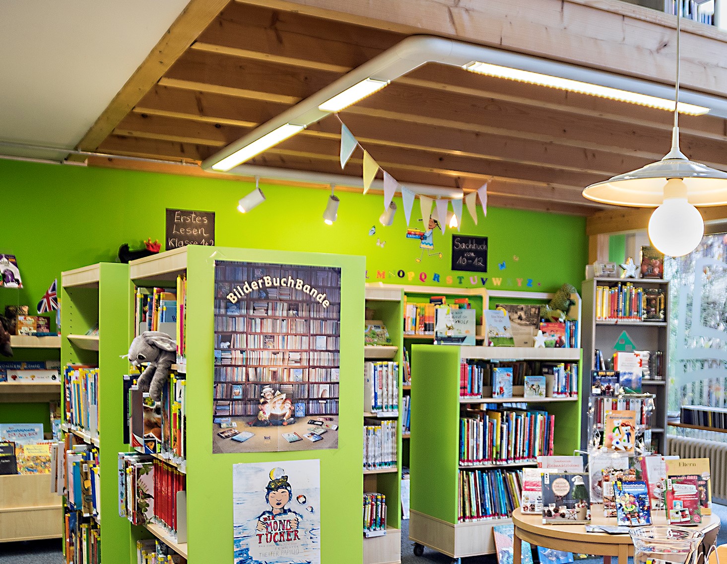 Das Bild zeigt einen Blick in die Bücherei. Die Wände sind grün gestrichen. Grüne Regale stehen vor der Wand und auch als Raumteiler. An einem Regal hängen Poster und die Regale sind vollbestückt mit Büchern. Auf dem Bild sieht man auf der rechten Seite einen runden Tisch, ebenfalls mit Büchern bedeckt. Von der Decke hängen Girlanden sowie eine weiße Schirmlampe herunter.