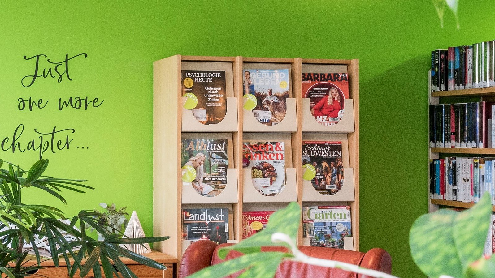 Auf dem Bild ist eine grüne Wand mit einem Tattoo das heißt, Just one more chapter ... zu sehen. Vor der grünen Wand steht ein Regal mit verschiedenen Zeitschriften darin. Auf der linken Seite vom Bild ist ebenfalls ein Regal mit vielen Büchern zu sehen. Im Vordergrund stehen grüne Pflanzen und ganz leicht ist ein roter Sessel ebenfalls im Vordergrund zu sehen.