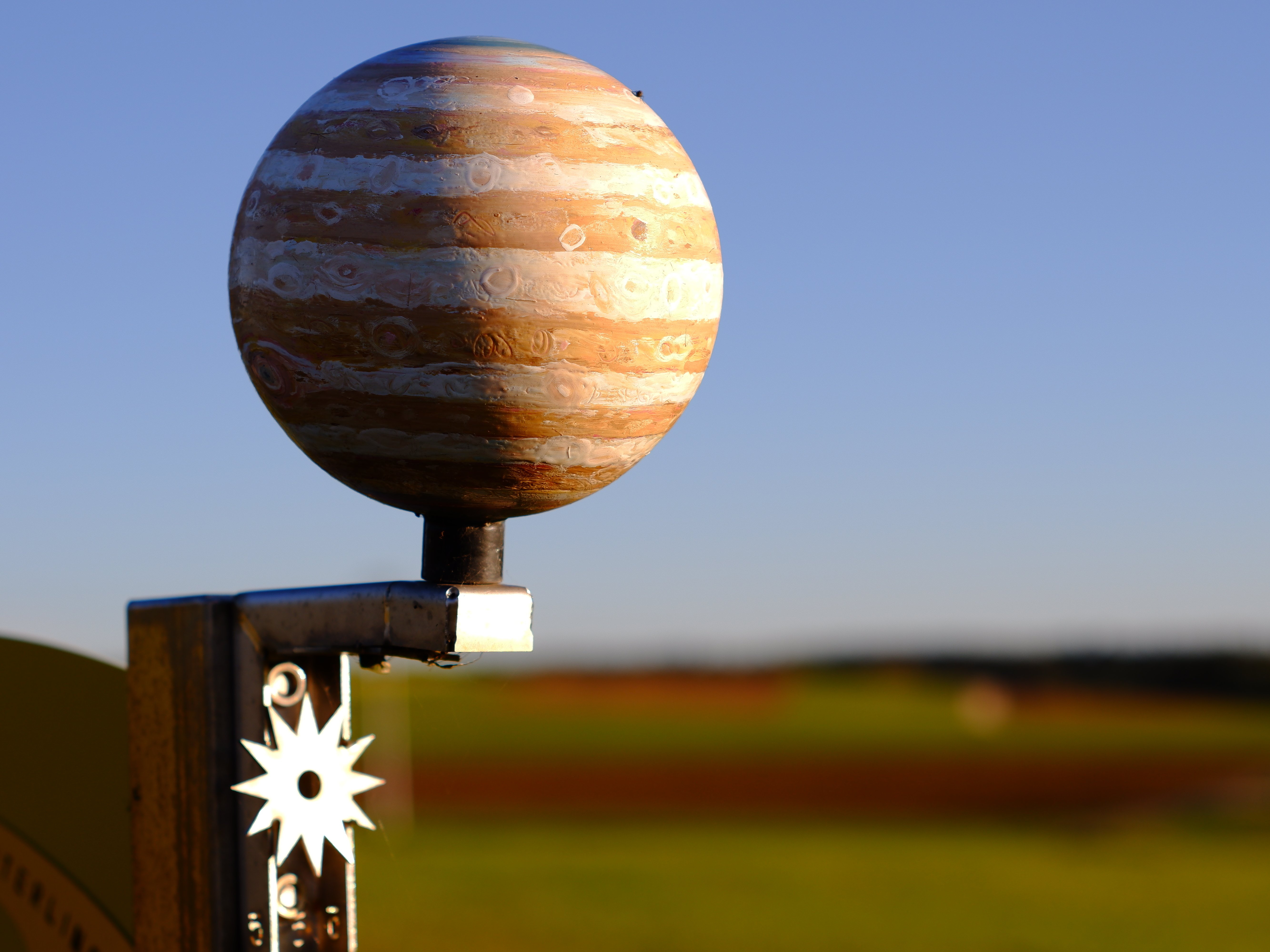 Auf dem Bild ist der Planet Jupiter in seiner hölzernen Farbe zu sehen. Im Hintergrund verschwommen der blaue Himmel und die grüne Wiese