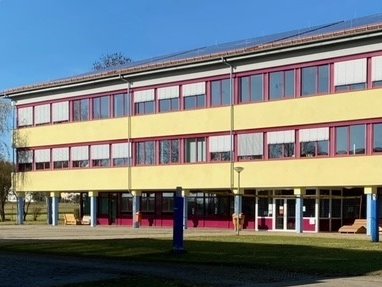 Auf dem Bild zeigt es die Realschule Winterlingen. Die Farbe der Realschule ist gelb und die Fensterrahmen sind rot. Auf dem Dach ist leicht die Photovoltaikanlage zu sehen. Der Himmel ist auf diesem Bild blau.